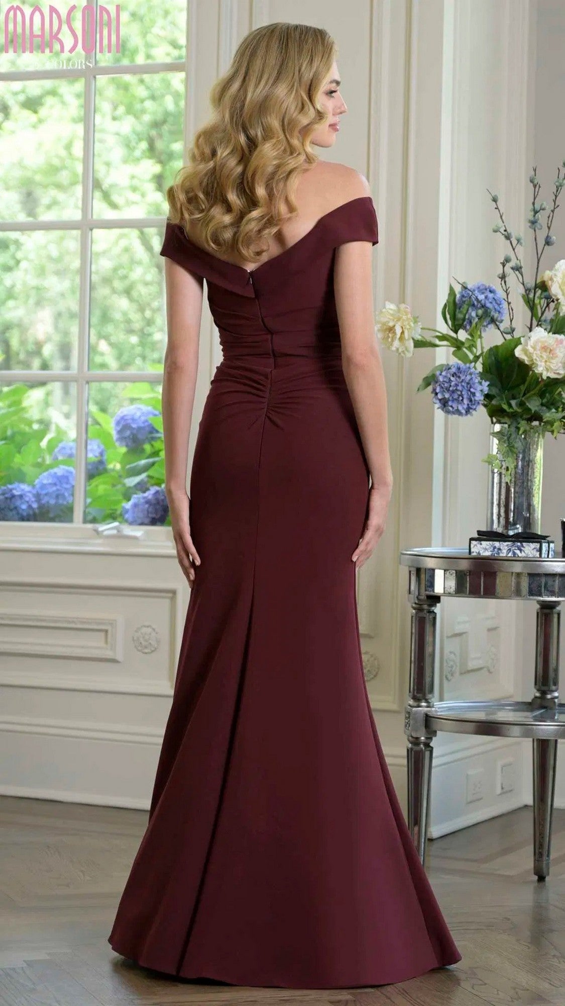 A woman with long wavy blonde hair wears the Marsoni M342 long formal dress in floor-length burgundy stretch crepe, standing indoors and facing away from the camera.